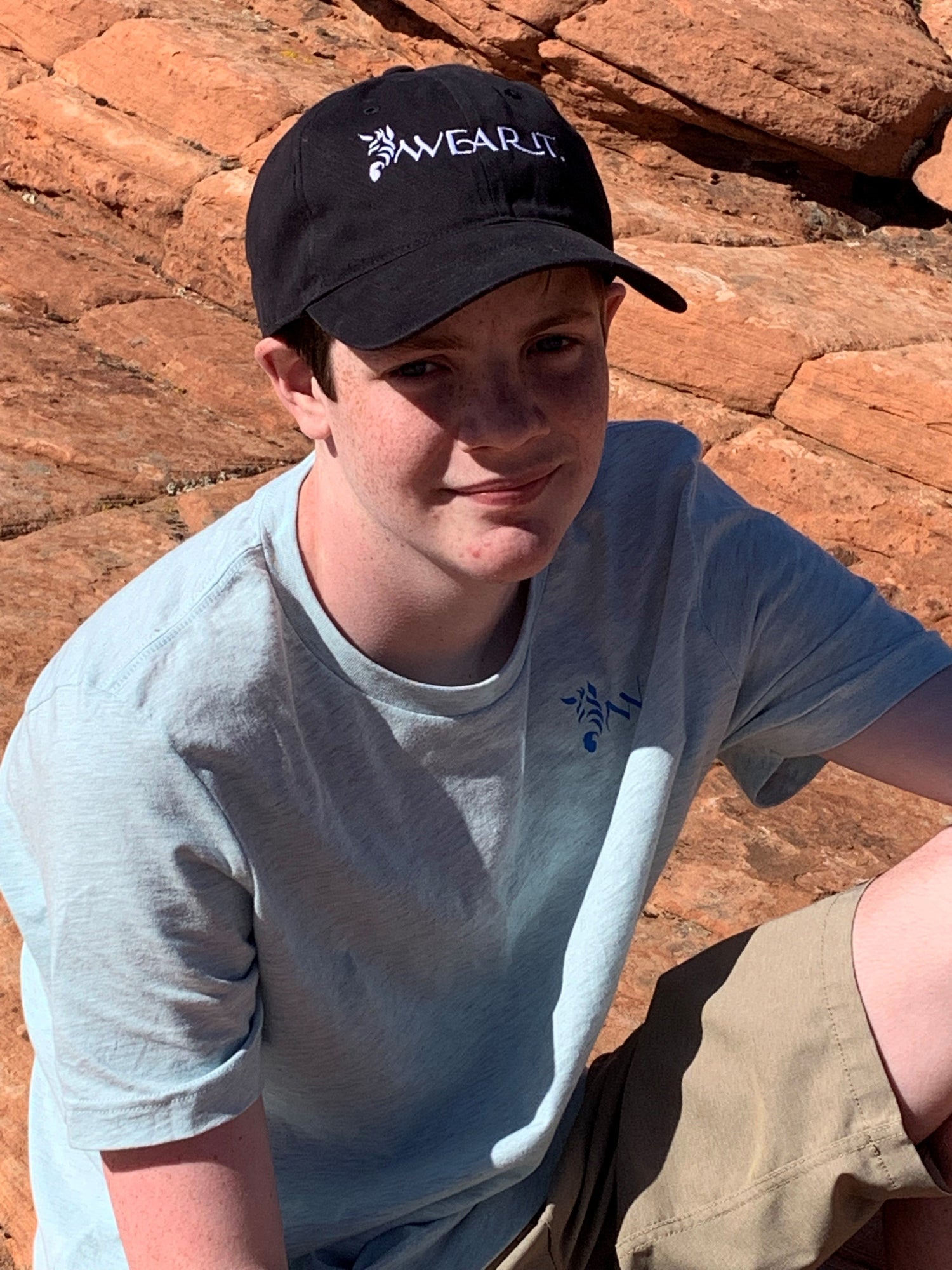 Person wearing a light blue WearIt. unisex t-shirt and black WearIt. baseball cap seated outdoors on red rock terrain