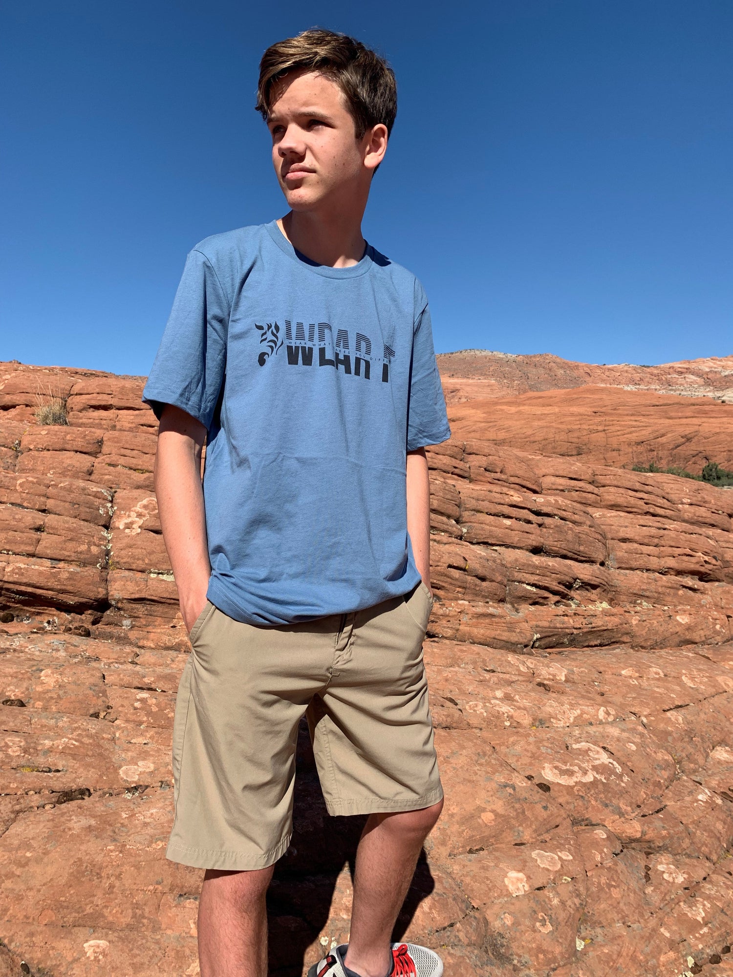 Person wearing a blue WearIt. unisex t-shirt standing outdoors on red rock terrain under a clear blue sky